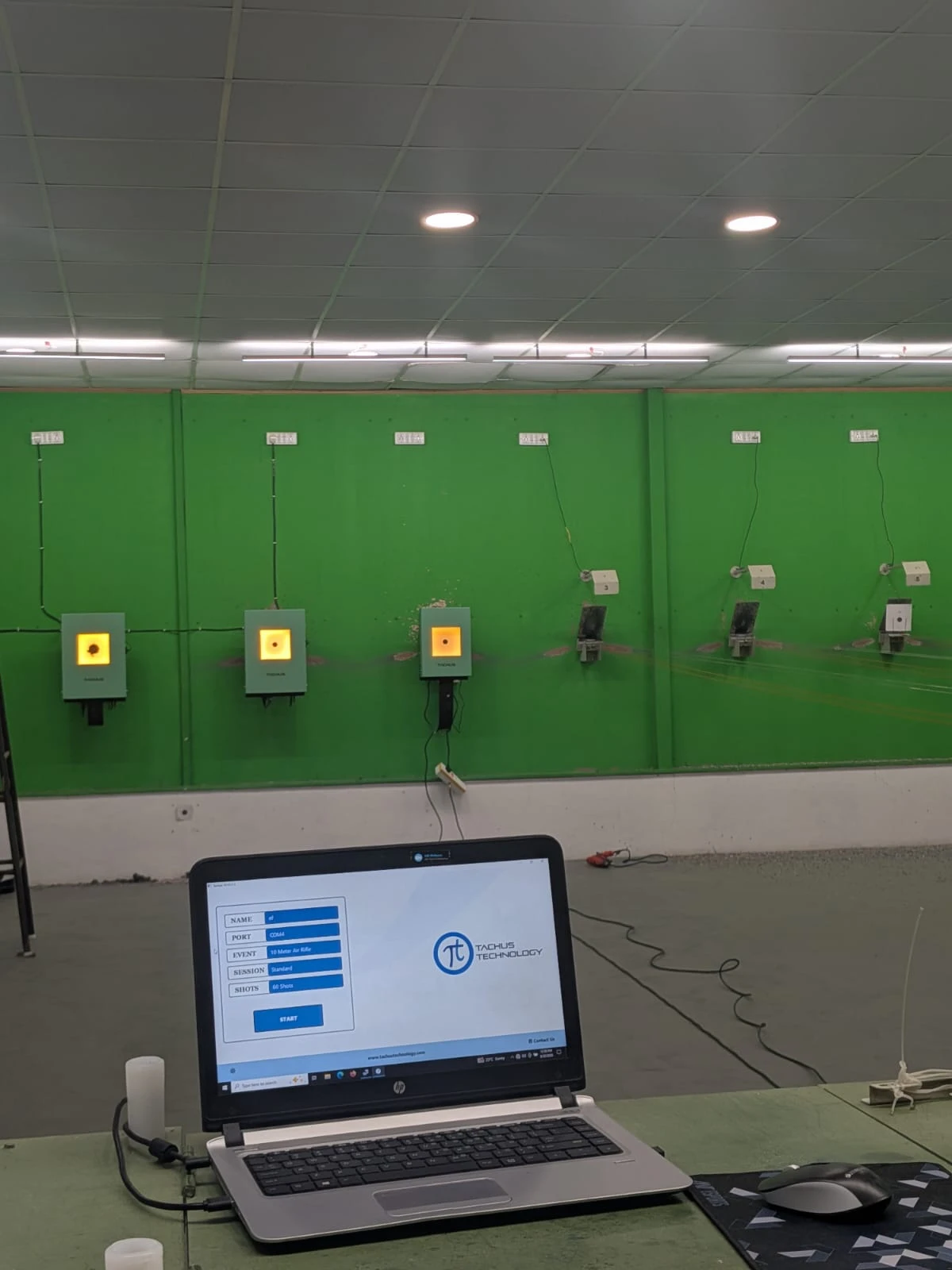 Tachus electronic targets on a green indoor range wall with a scoring laptop in the foreground